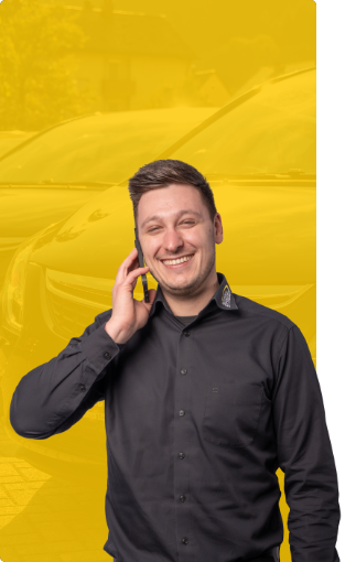 Ein lächelnder Mann in einem schwarzen Hemd telefoniert mit einem Mobiltelefon und steht vor gelb getönten Autos.