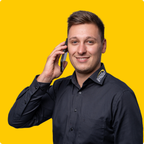 Ein junger Mann in einem schwarzen Hemd lächelt, während er ein Smartphone an sein Ohr hält, und steht vor einem leuchtend gelben Hintergrund.