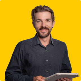 Ein Mann mit braunen Haaren und Bart, der ein dunkles Button-up-Hemd trägt, lächelt, während er ein digitales Tablet vor einem hellgelben Hintergrund hält.